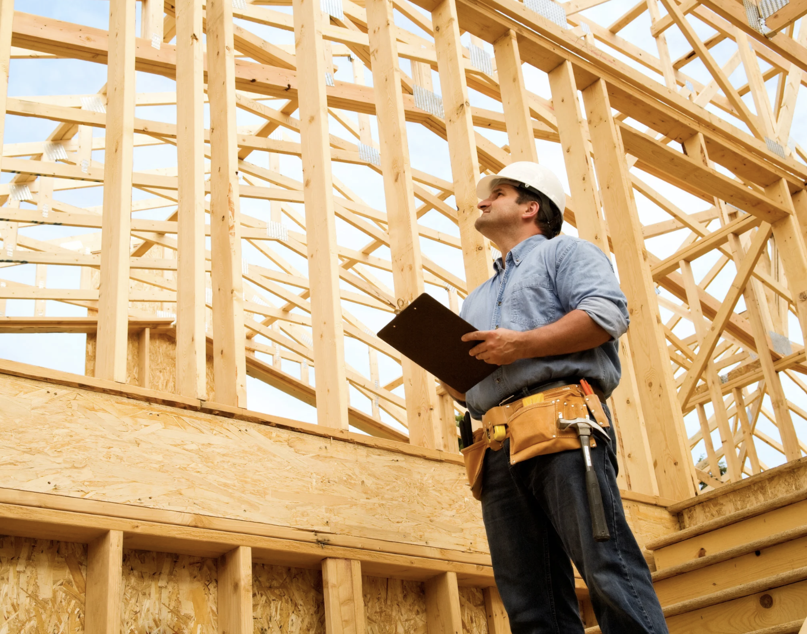 Construction worker in hard hat and safety vest standing inside wooden framework of building under construction, looking at a tablet